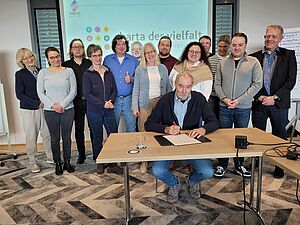 Große Gruppe von Menschen stehend hinter an einem Tisch. Am Tisch sitzt ein Mann und unterschreibt ein Dokument.